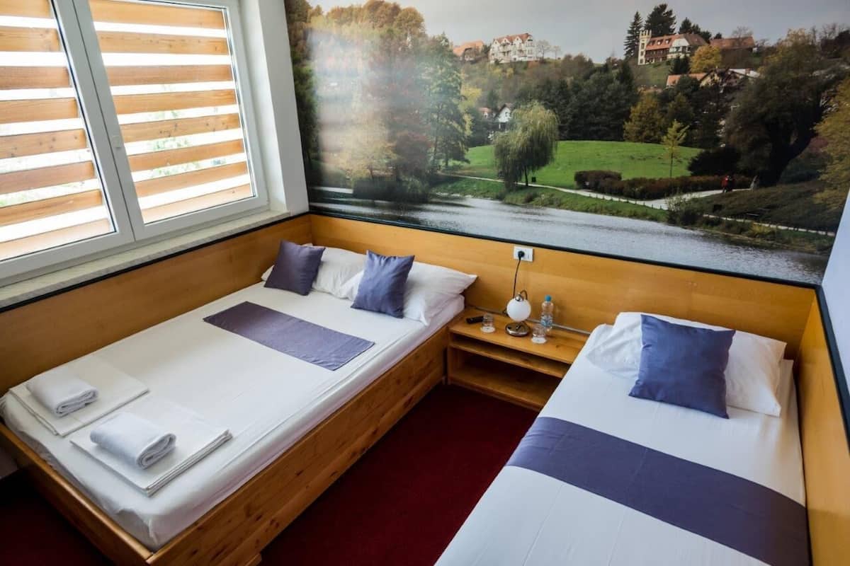 A hotel room with two beds, a wooden headboard, a large window with blinds, and a view of a landscape with trees and a building.