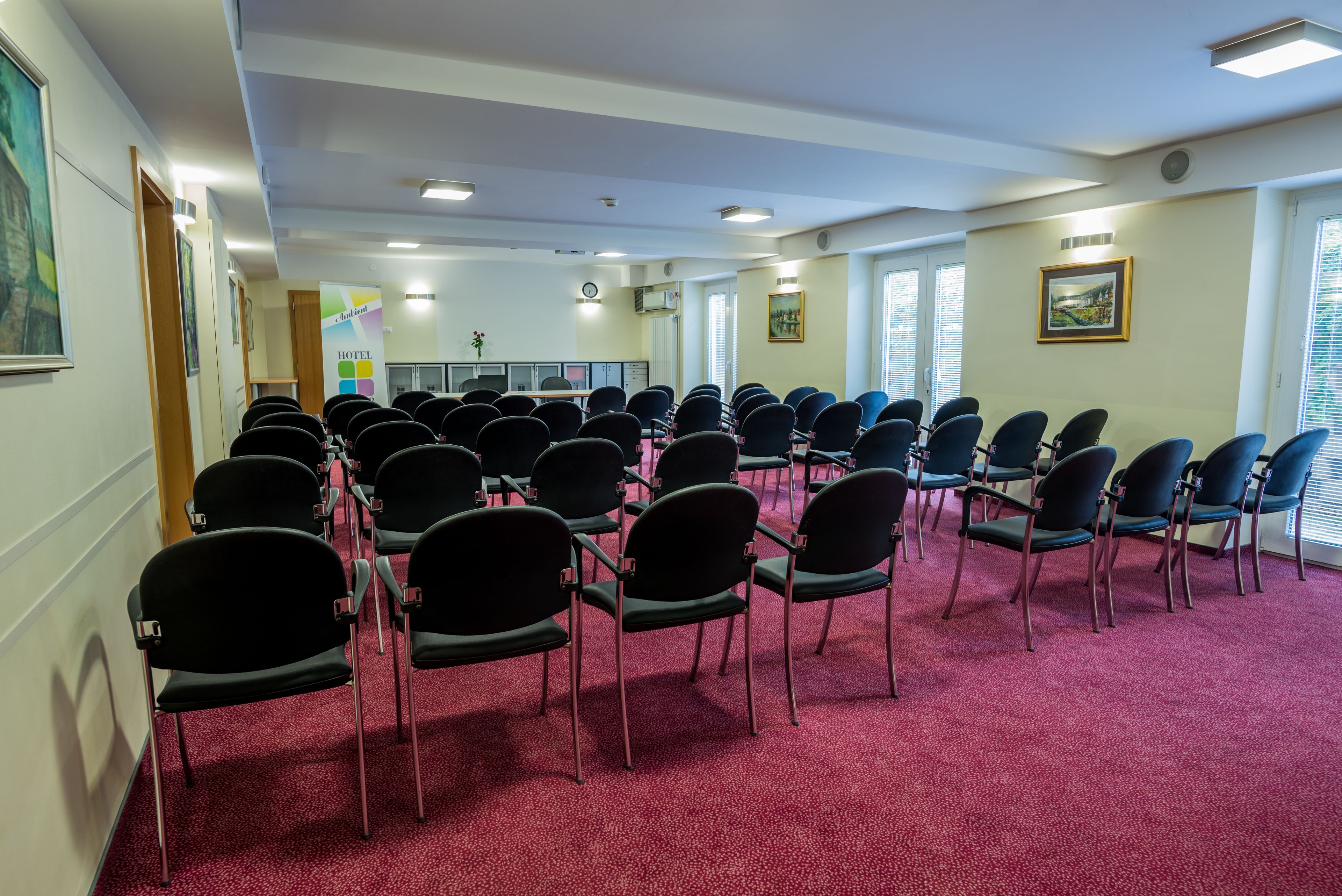 Uma sala de conferências com fileiras de cadeiras, piso coberto por carpete e obras de arte emolduradas nas paredes.