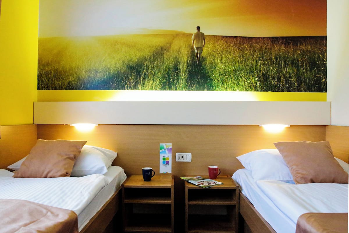 A hotel room with two beds, each with a pillow and a nightstand. There is a painting of a field on the wall above the beds.