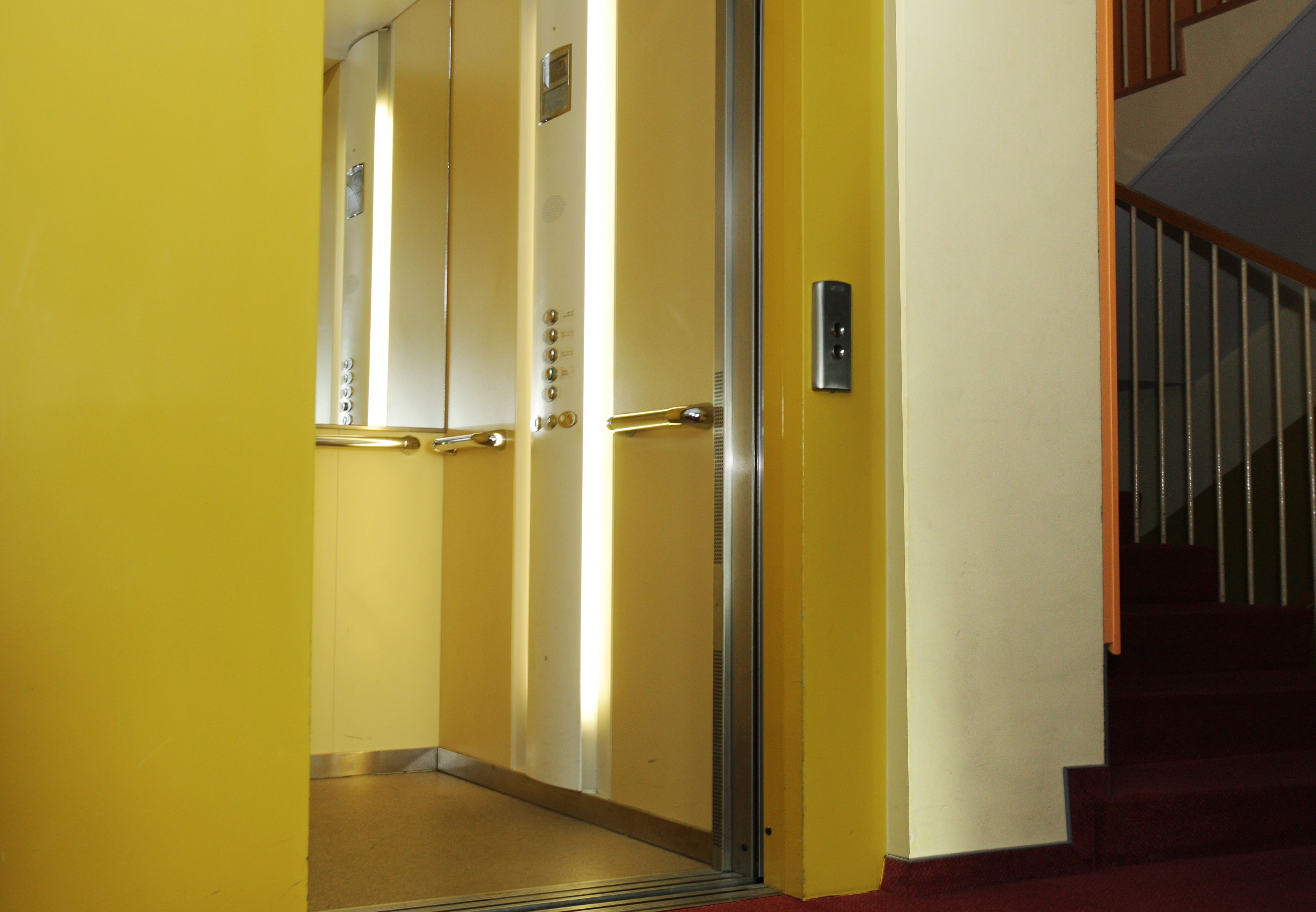 An elevator with metallic doors and a control panel, leading to a staircase with wooden railings.