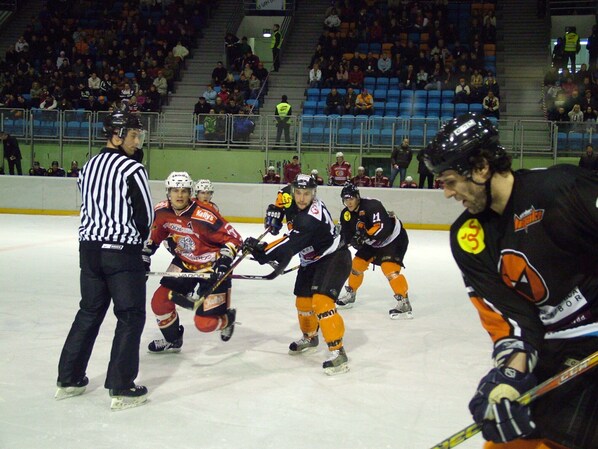 An indoor ice hockey rink with players in red and black jerseys, a referee, and spectators in the background.