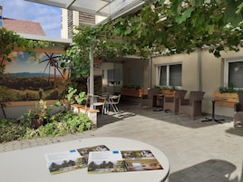 Un espacio al aire libre con mesas y sillas, rodeado de vegetación y un mural con un paisaje.