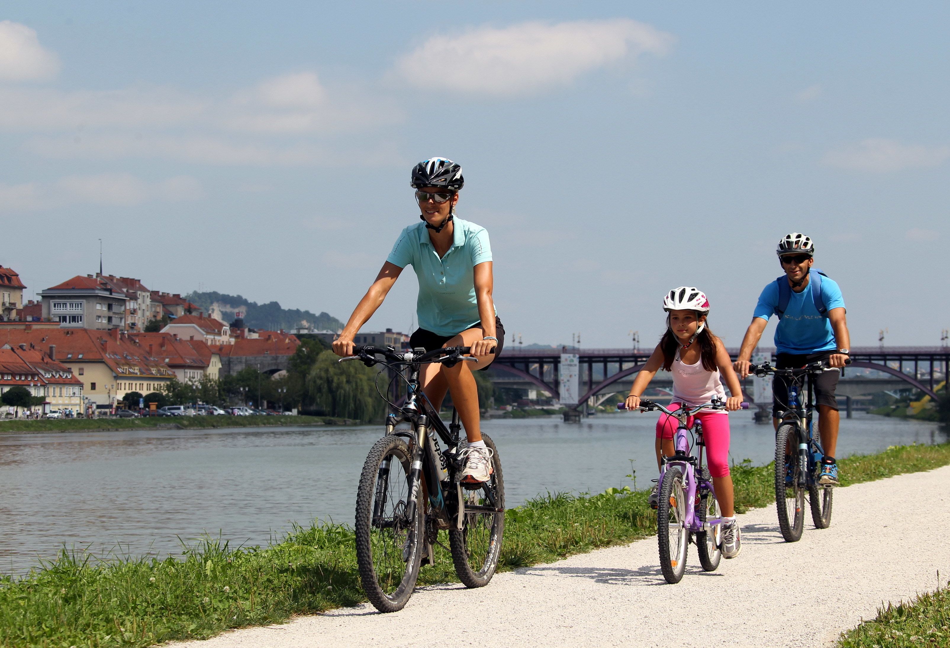 Tres personas andando en bicicleta por un sendero junto a un río, con edificios y un puente al fondo.