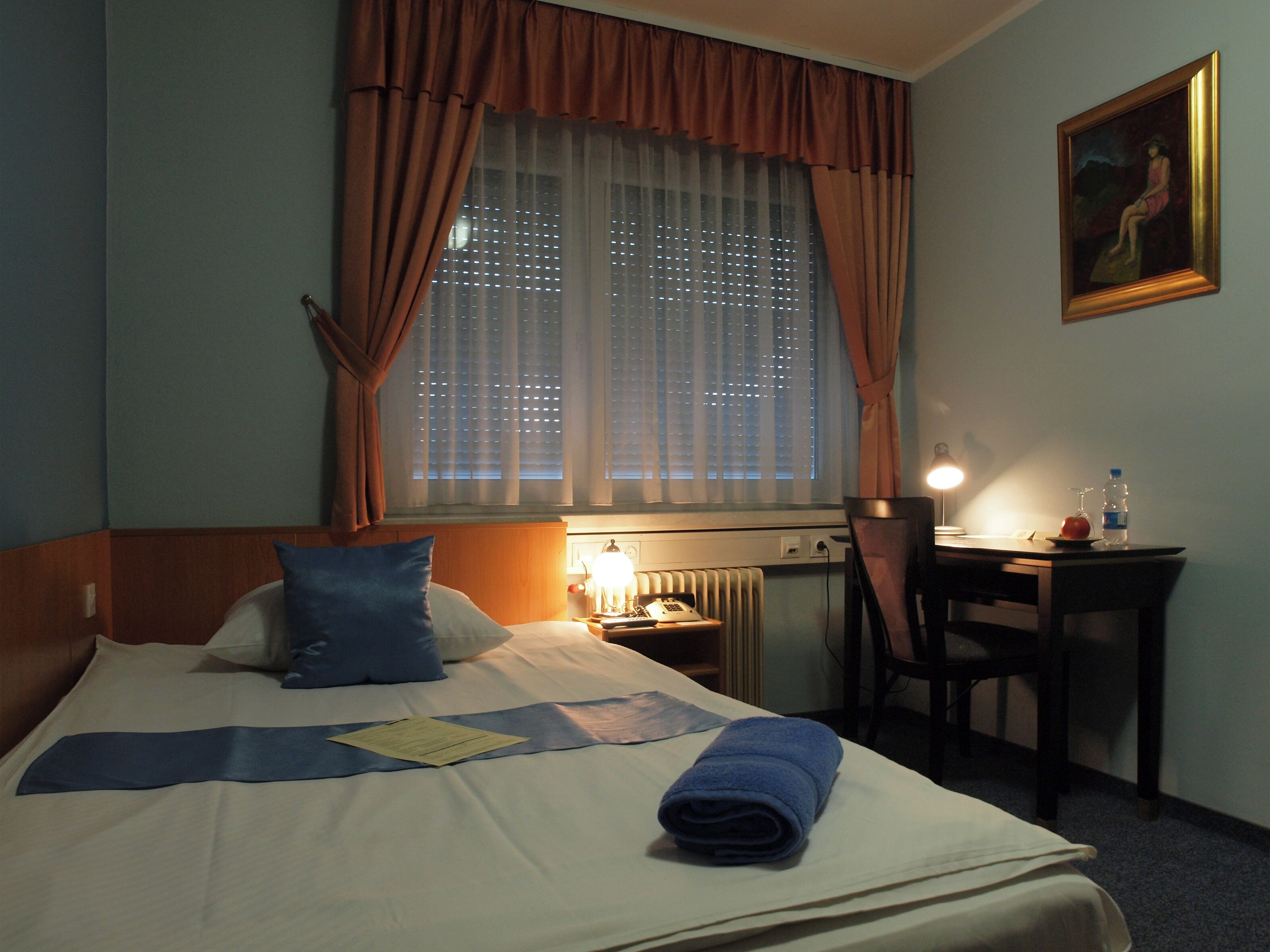 Habitación de hotel con cama, escritorio, silla, lámpara y un cuadro en la pared.