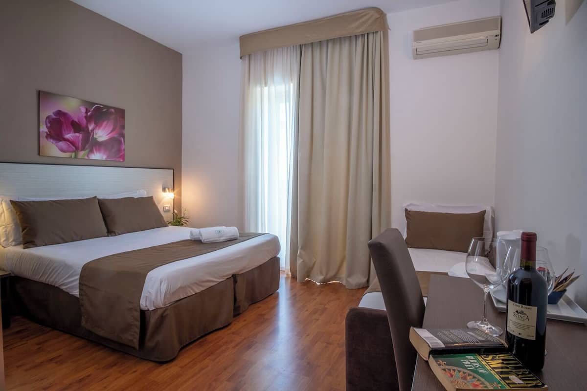 A hotel room with a bed, a desk with a bottle of wine and glasses, a chair, and a window with curtains.