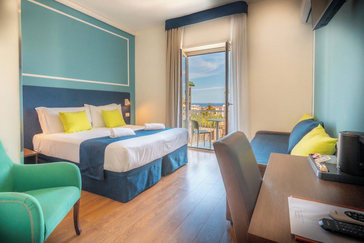 A hotel room with a bed, two chairs, a desk, and a view of the outside.