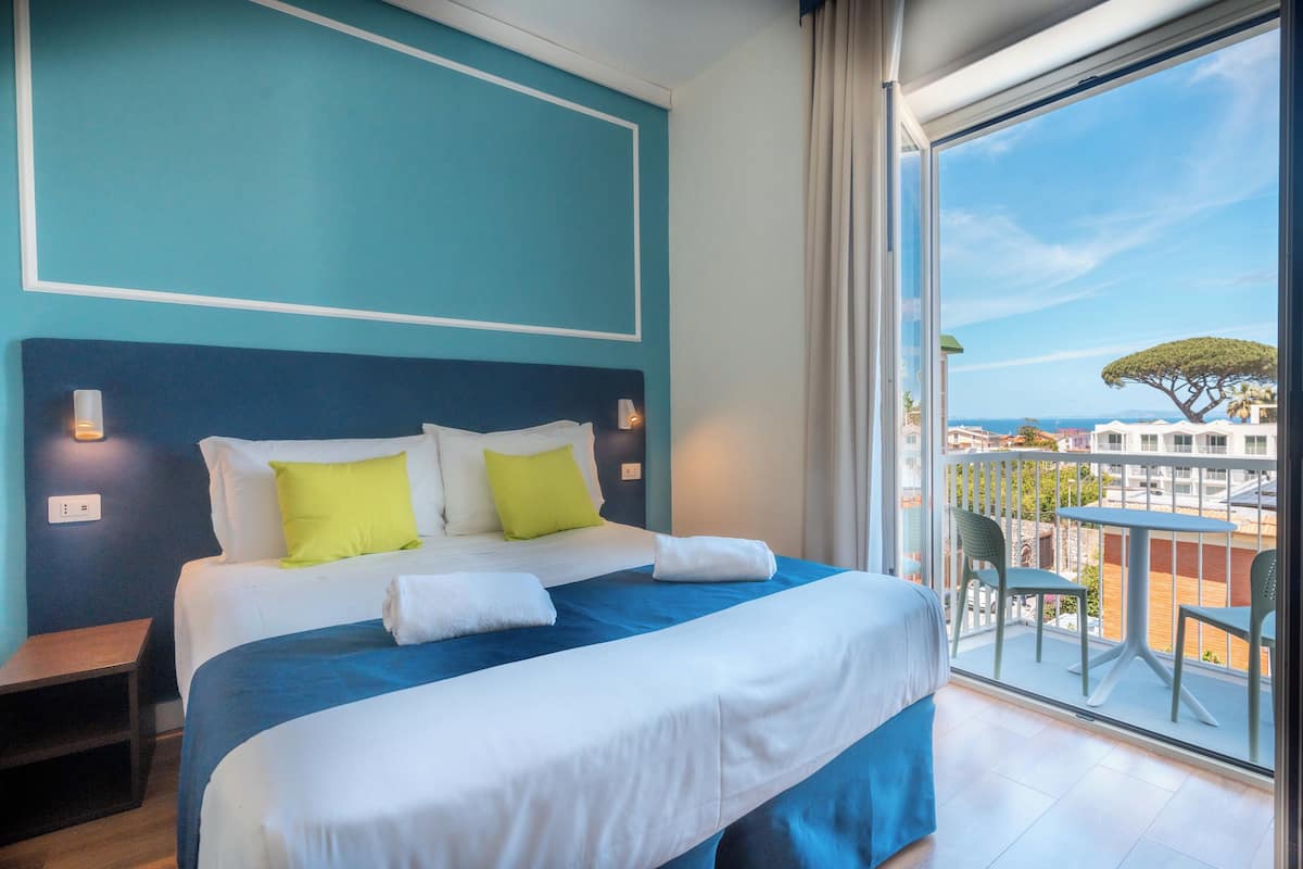 A hotel room with a large bed, two pillows, a nightstand, and a balcony with a table and chairs.