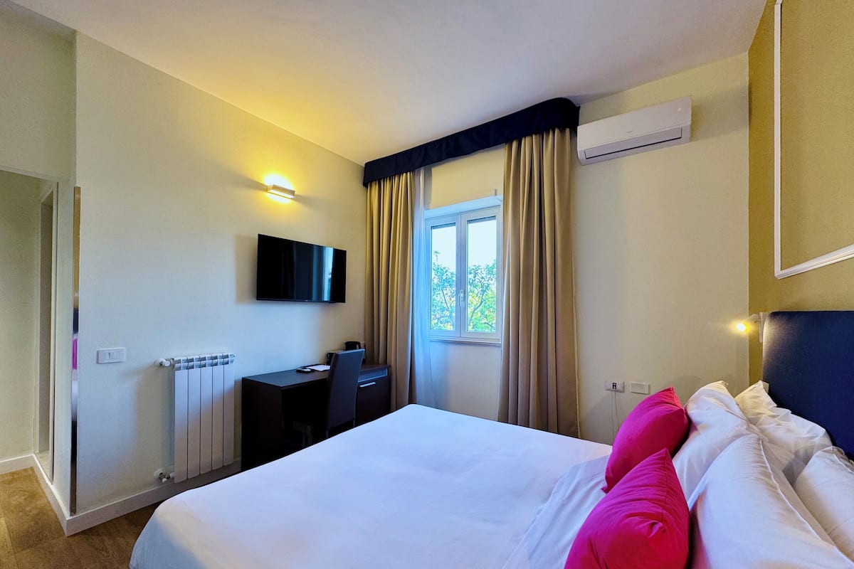 A hotel room with a bed, a desk, a chair, a TV, and an air conditioning unit.