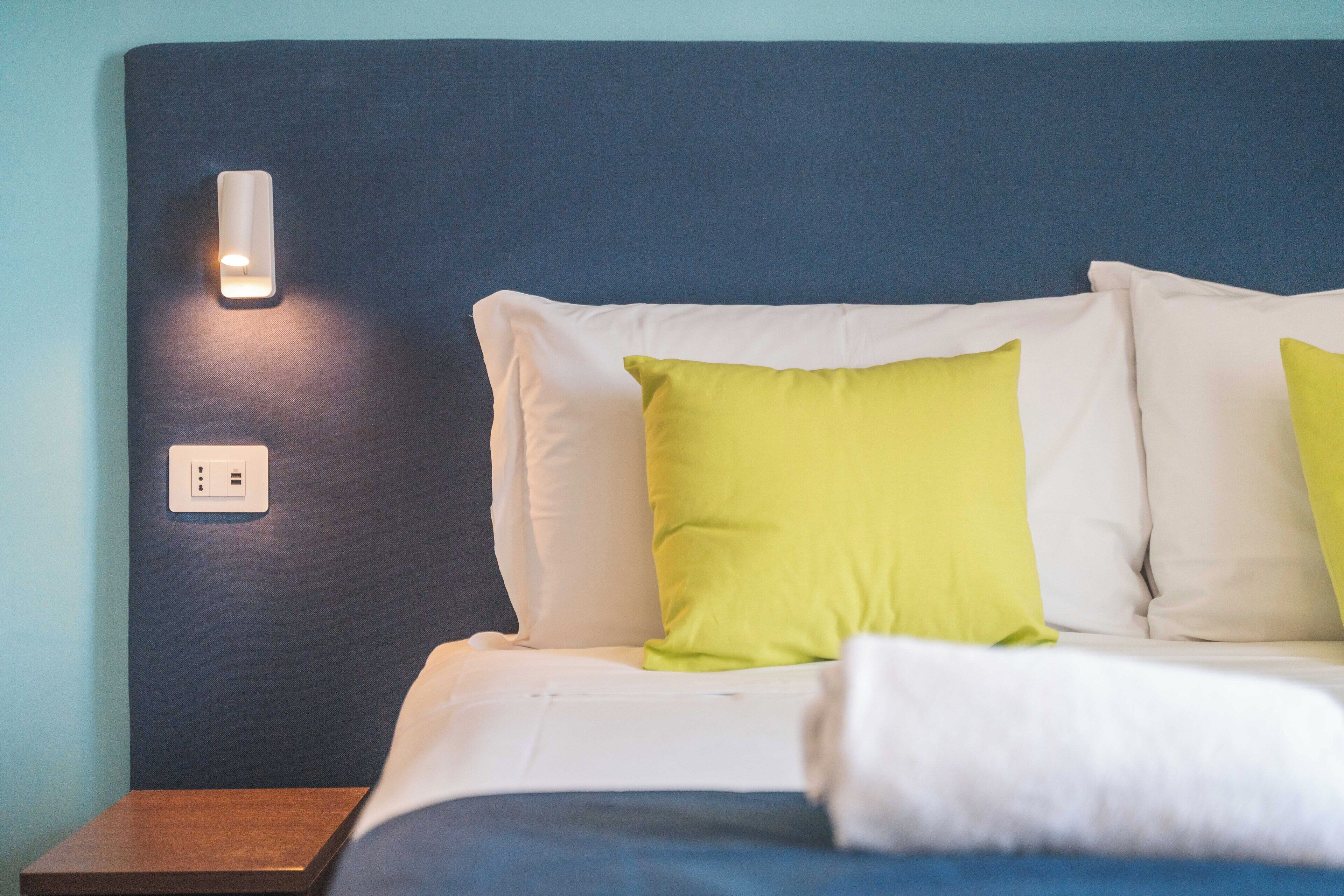 A neatly made bed with white and green pillows, a wooden nightstand, and a wall-mounted light fixture.
