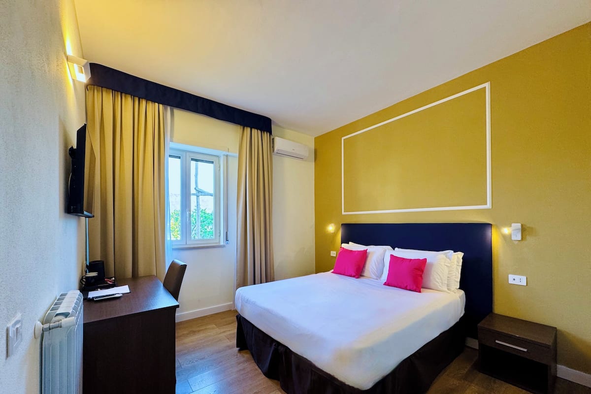 A hotel room with a bed, a desk, a chair, a TV mounted on the wall, and a window with curtains.