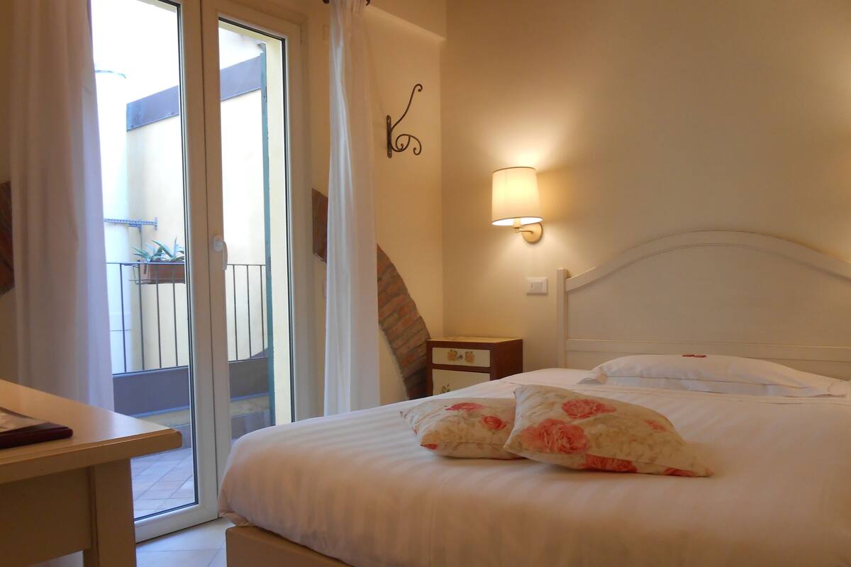 A hotel room with a bed, a nightstand, a wall-mounted lamp, a sliding glass door to a balcony, and a small table.