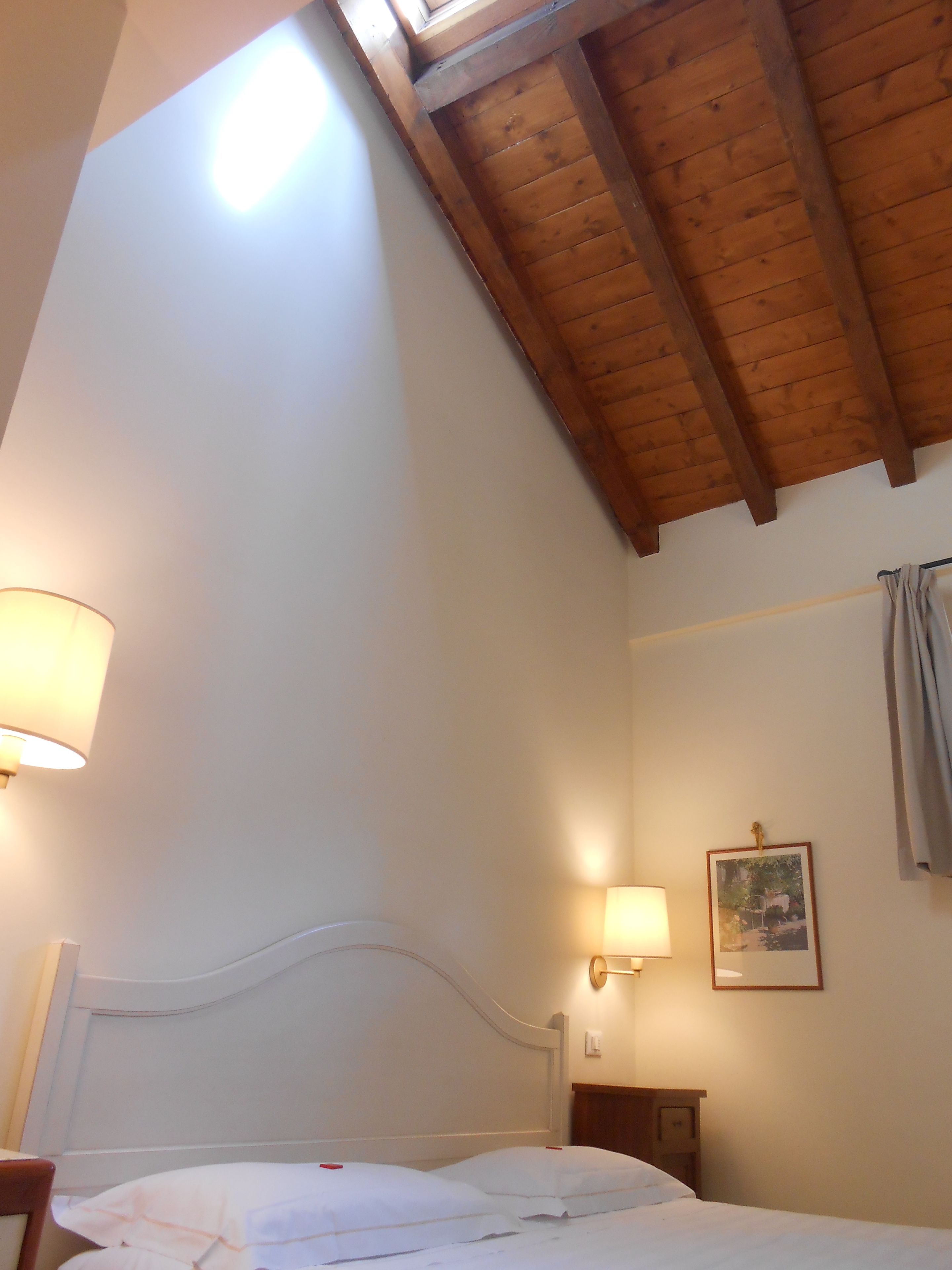 A bedroom with a wooden ceiling, a bed, two wall-mounted lamps, and a framed picture on the wall.