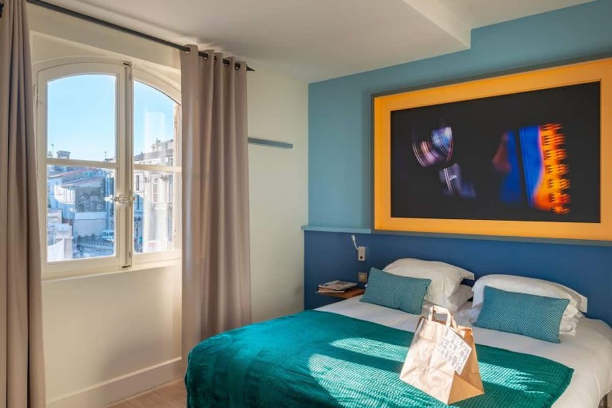 Una habitación de hotel con cama, ventana grande y un cuadro en la pared.
