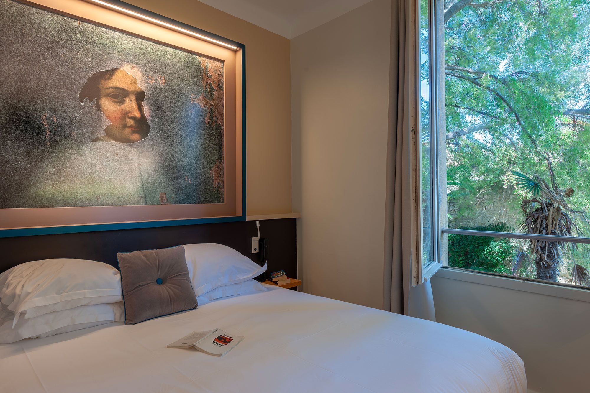 Ein Hotelzimmer mit einem großen Bett, einem gerahmten Kunstwerk darüber, einem Nachttisch mit einem Buch und einem Fenster mit Blick auf Bäume.