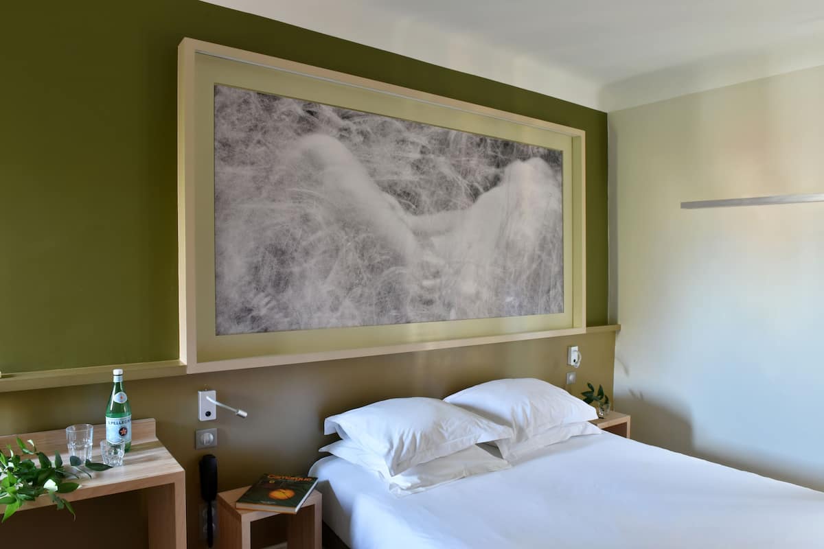 Habitación de hotel con cama, mesita de noche, un cuadro colgado en la pared, una botella de agua y un libro.