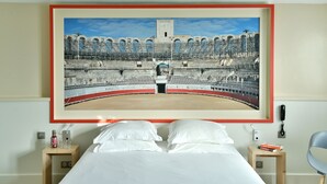 Une chambre d’hôtel avec une grande photographie encadrée représentant un amphithéâtre antique, un lit avec du linge de maison blanc, une table de chevet avec une bouteille et des verres, et une petite table de chevet avec des livres.
