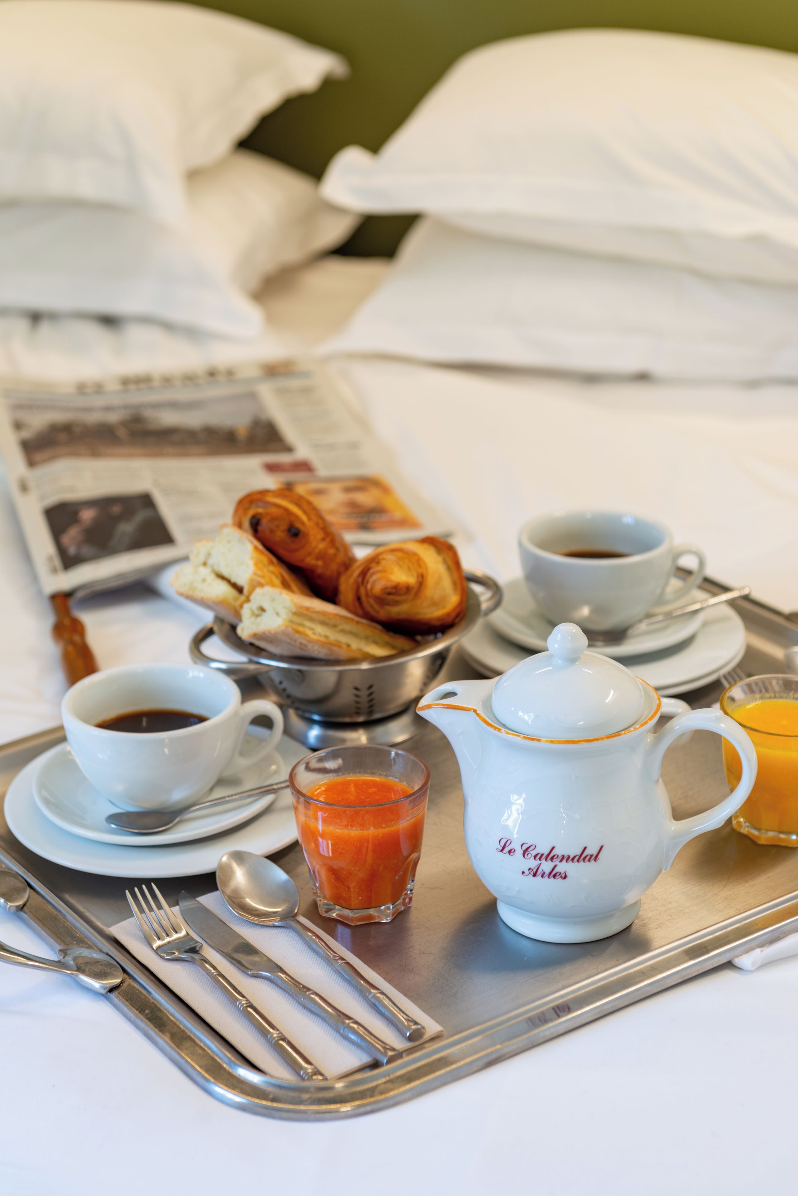 A breakfast tray with croissants, coffee, juice, and a teapot on a bed with pillows and a newspaper.