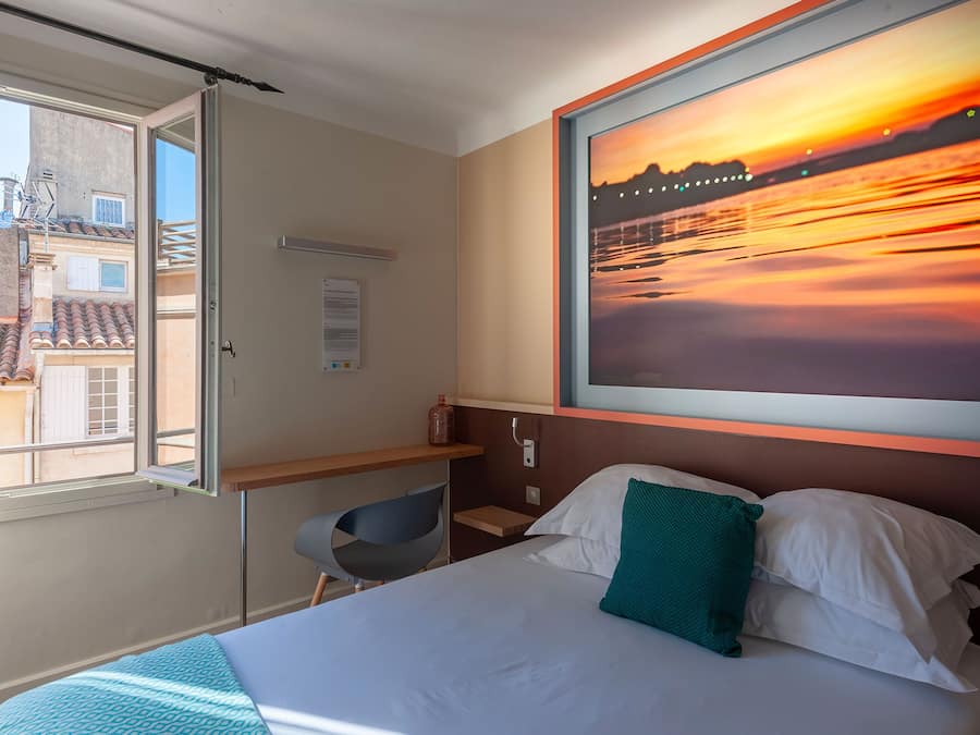 A hotel room with a bed, a desk, a chair, a window, and a sunset painting.