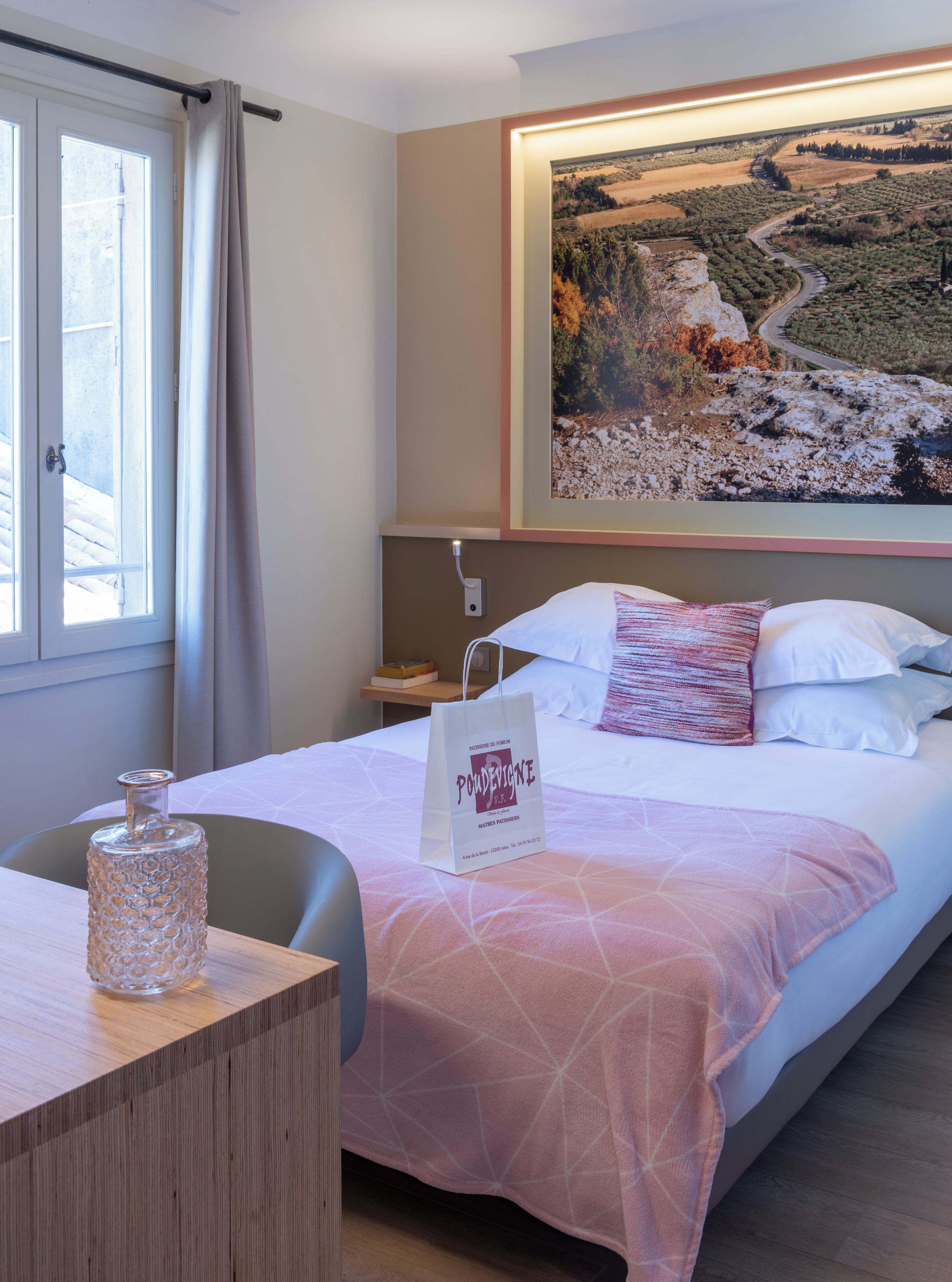 Une chambre d’hôtel avec un lit, un tableau au mur, une table de chevet avec un vase en verre et un sac posé sur le lit.