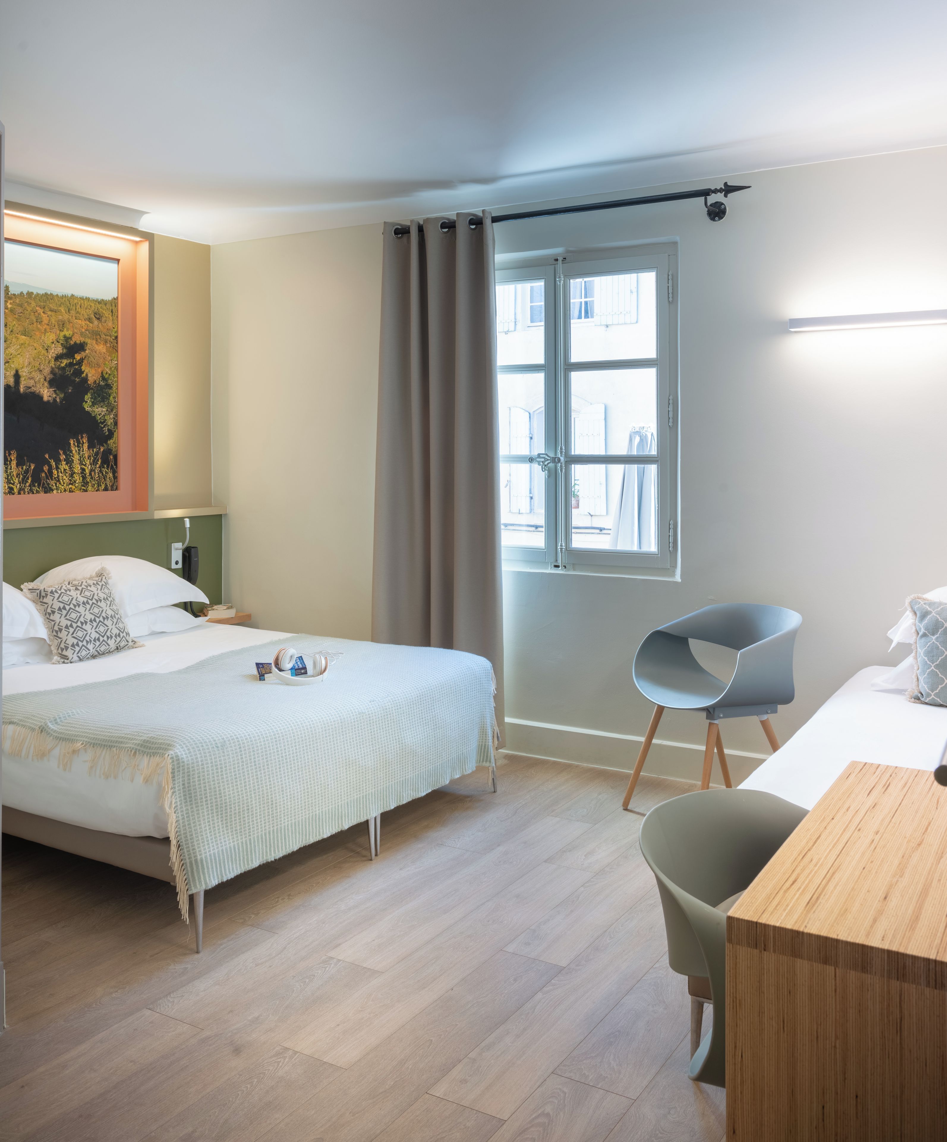 Ein Hotelzimmer mit Bett, Schreibtisch, Stuhl, einem Fenster mit Vorhängen und einem Bild an der Wand.