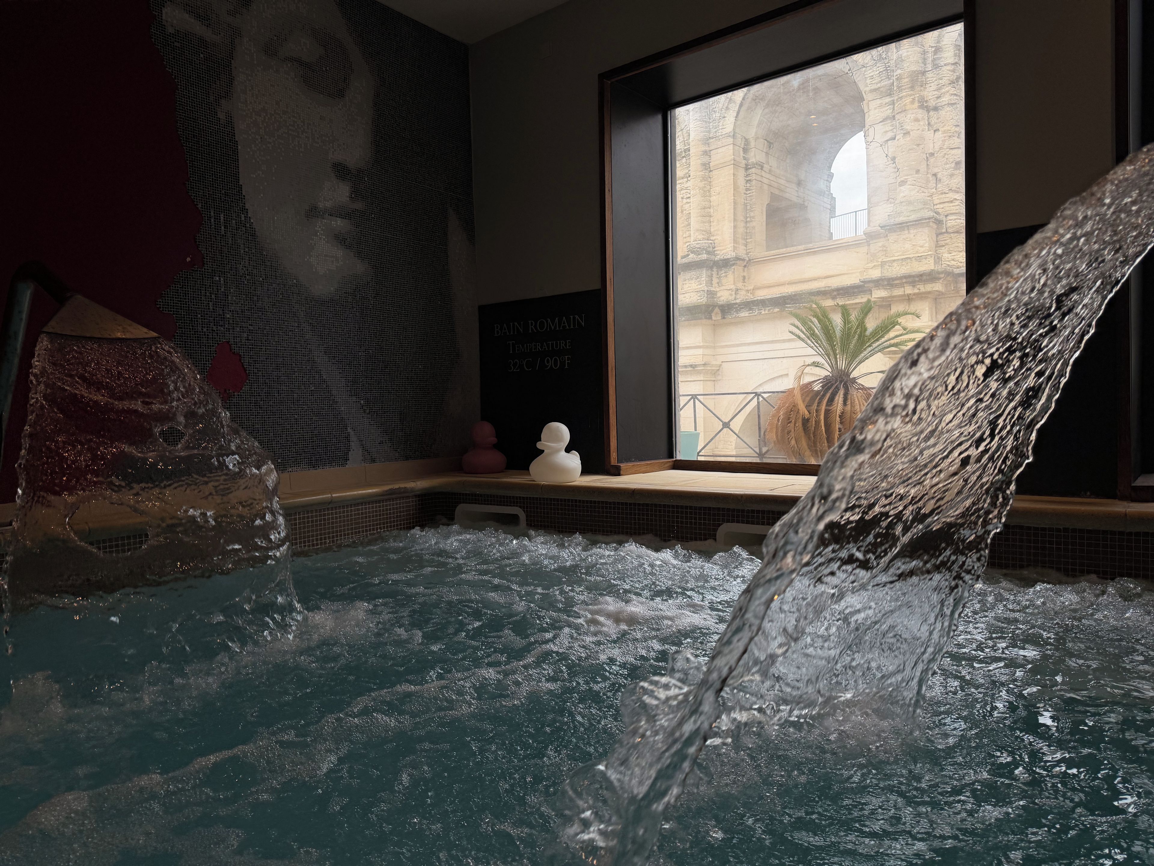 A hot tub with a waterfall feature, a mural, and a view of a building through a window.