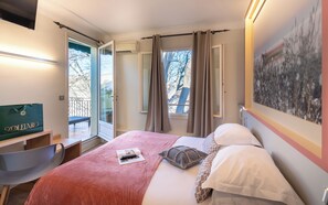 Una habitación de hotel con cama, escritorio, silla, ventana con cortinas y un cuadro en la pared.