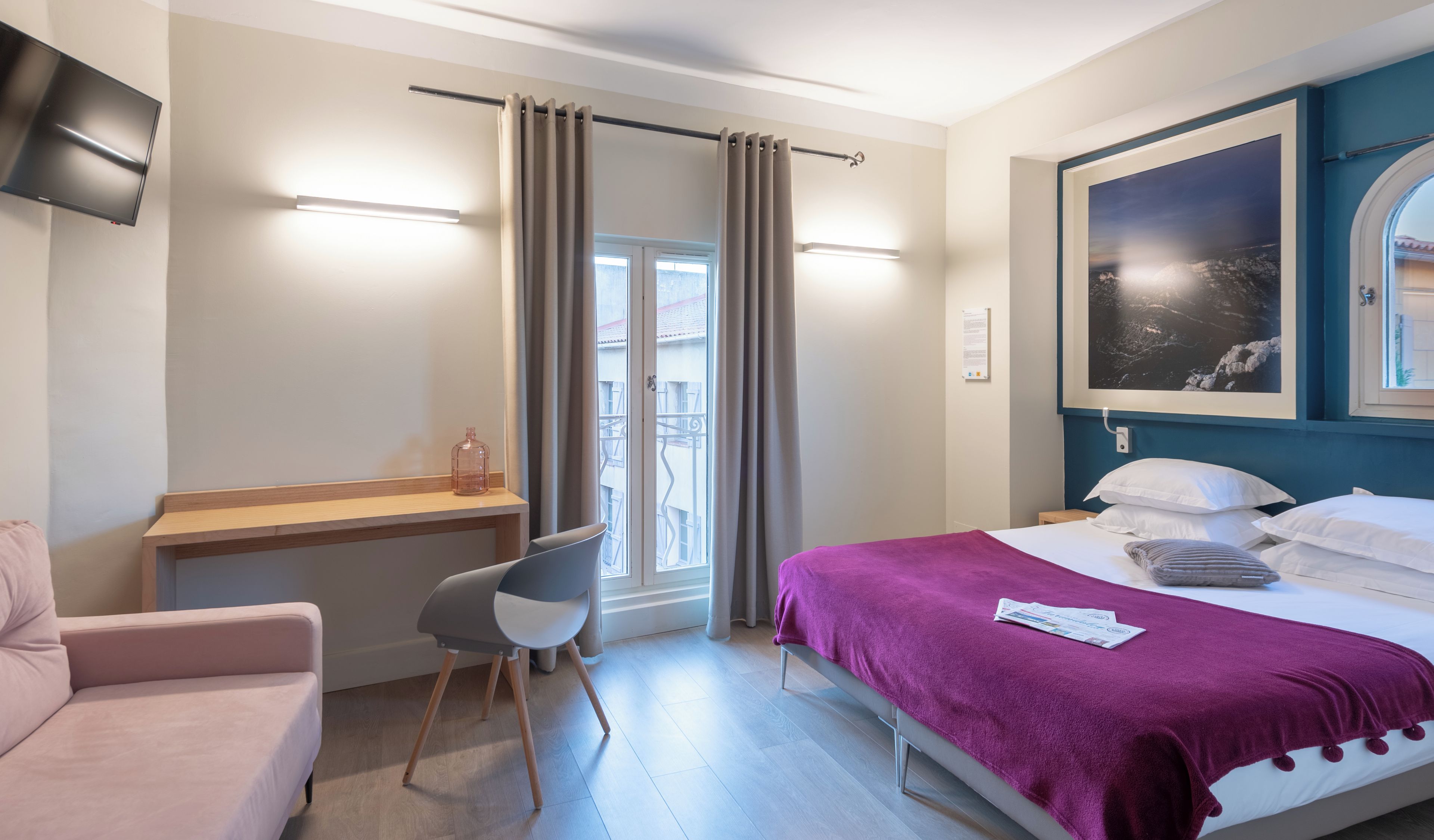 A hotel room with a bed, a desk, a chair, a TV, and a window with curtains.