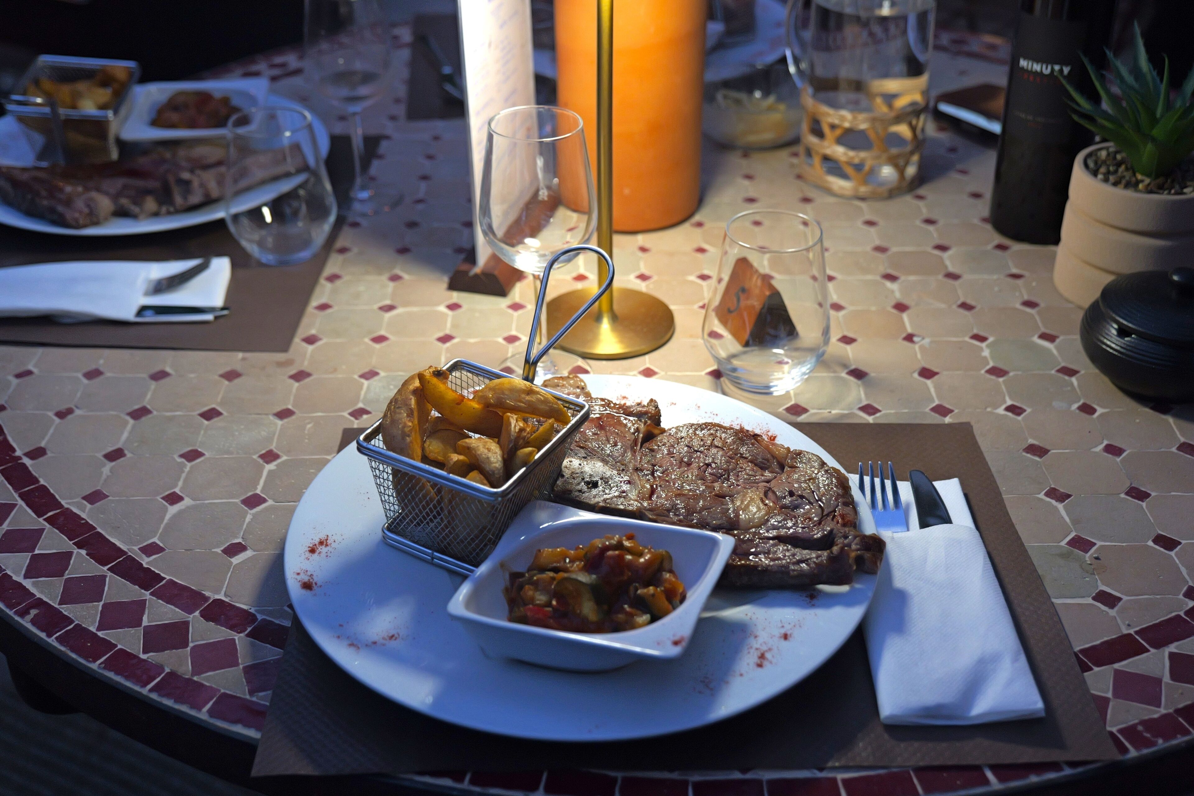 Una mesa de comedor con comida que incluye filete, papas fritas y un acompañamiento, además de una vela y una planta en maceta.