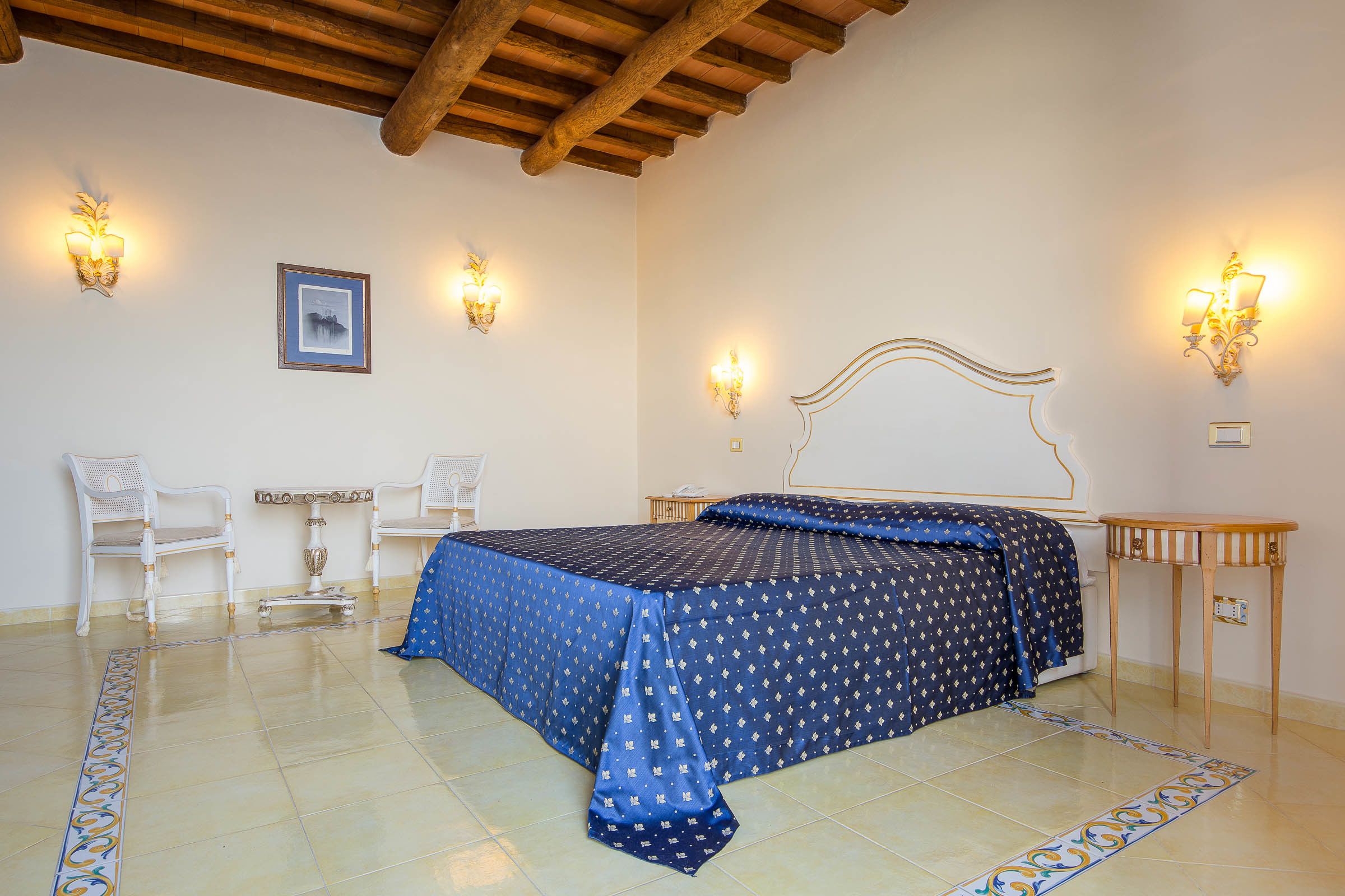 A bedroom with a bed, two chairs, a small table, and wall sconces.