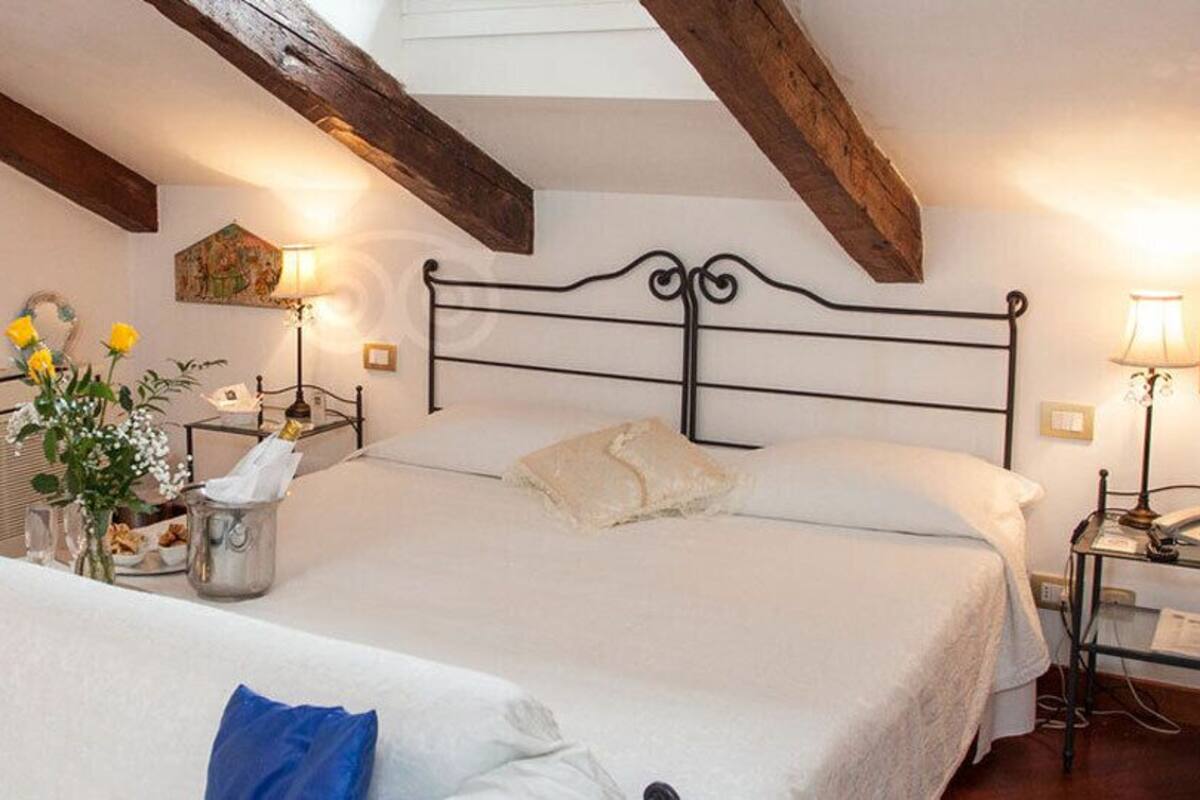 A bedroom with a large bed, a bedside table with a lamp, a small table with a vase of flowers, and a painting on the wall.