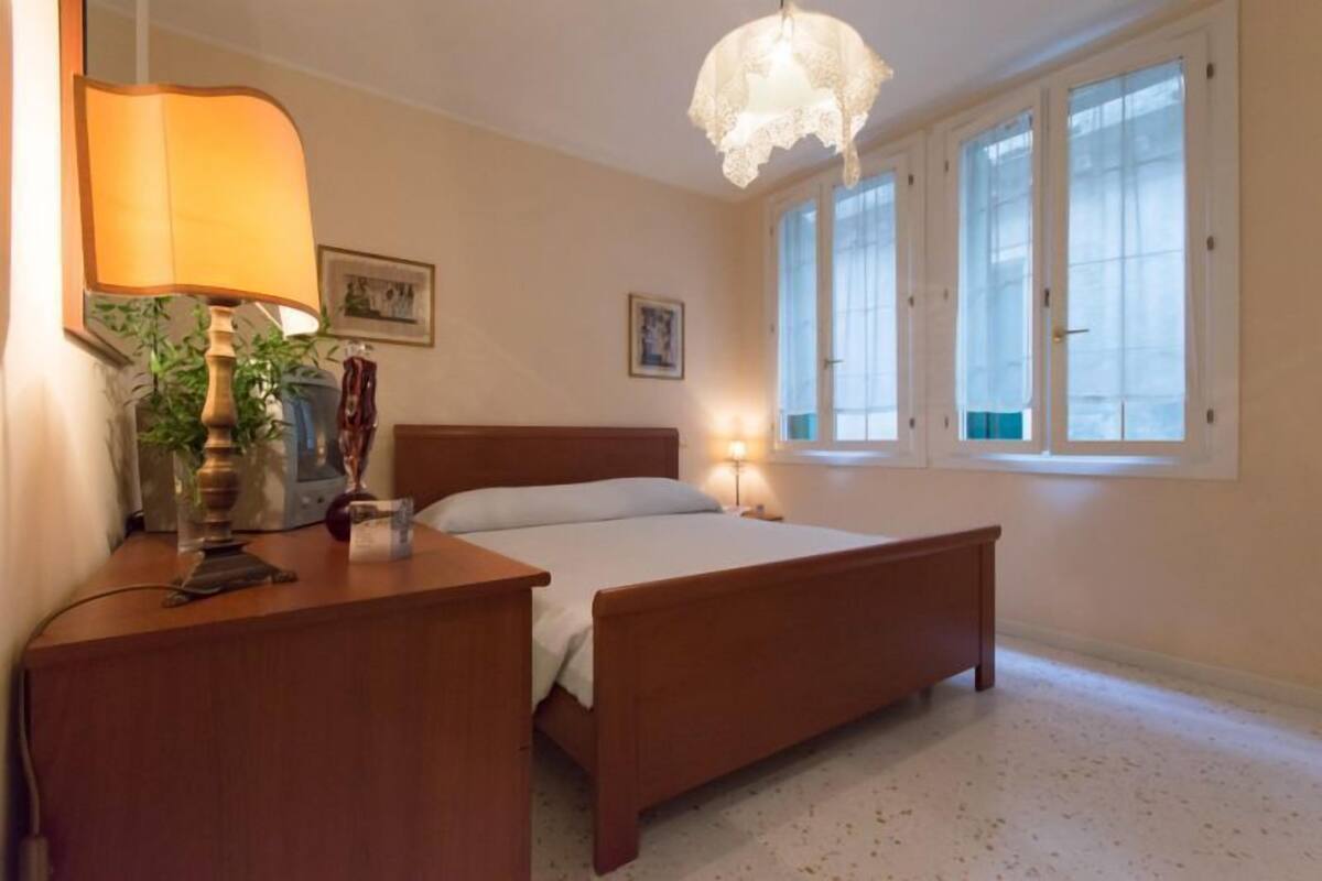 A bedroom with a wooden bed, a bedside table with a lamp, and a window with frosted glass.