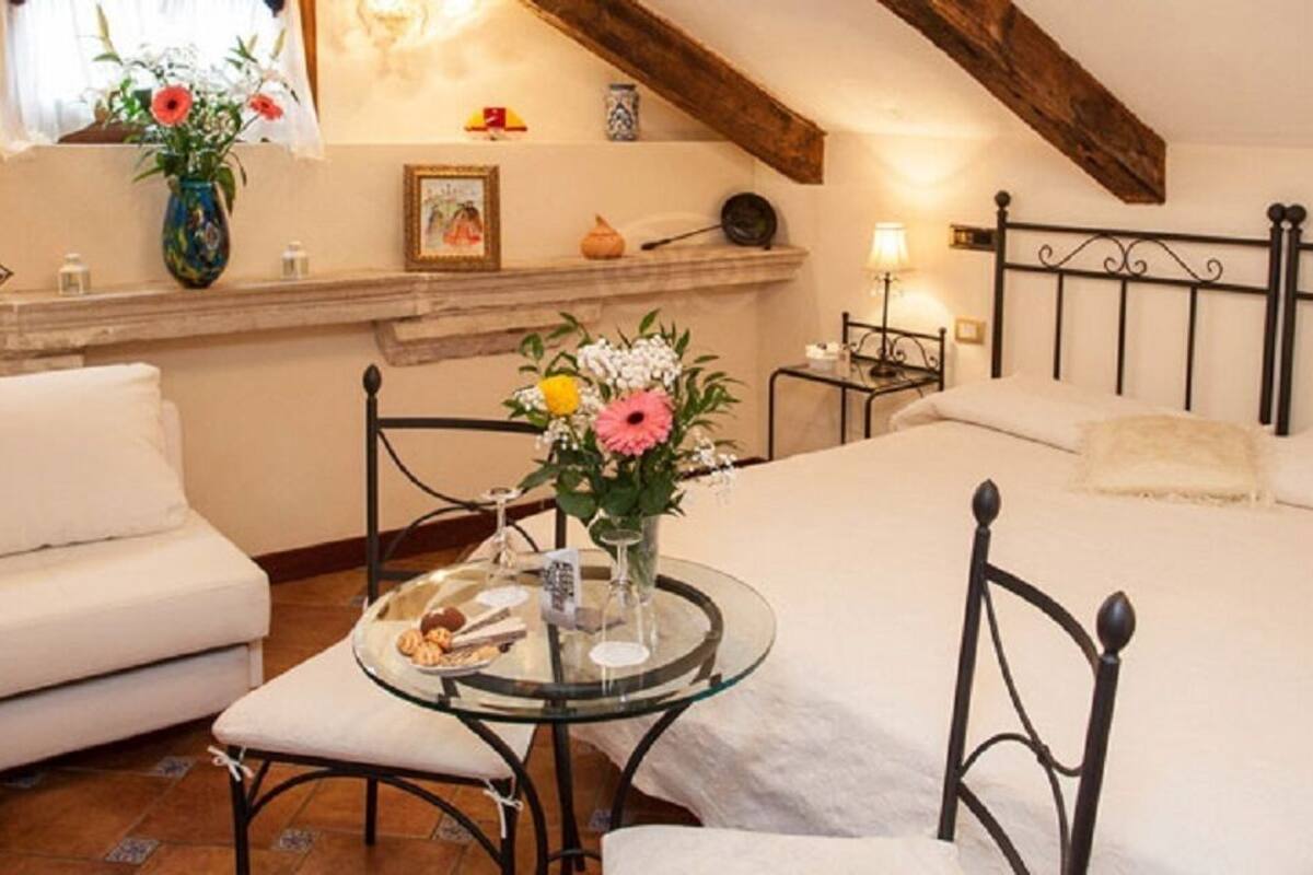 A cozy bedroom with a large bed, a sofa, a small round table with a vase of flowers, and a wooden ceiling.