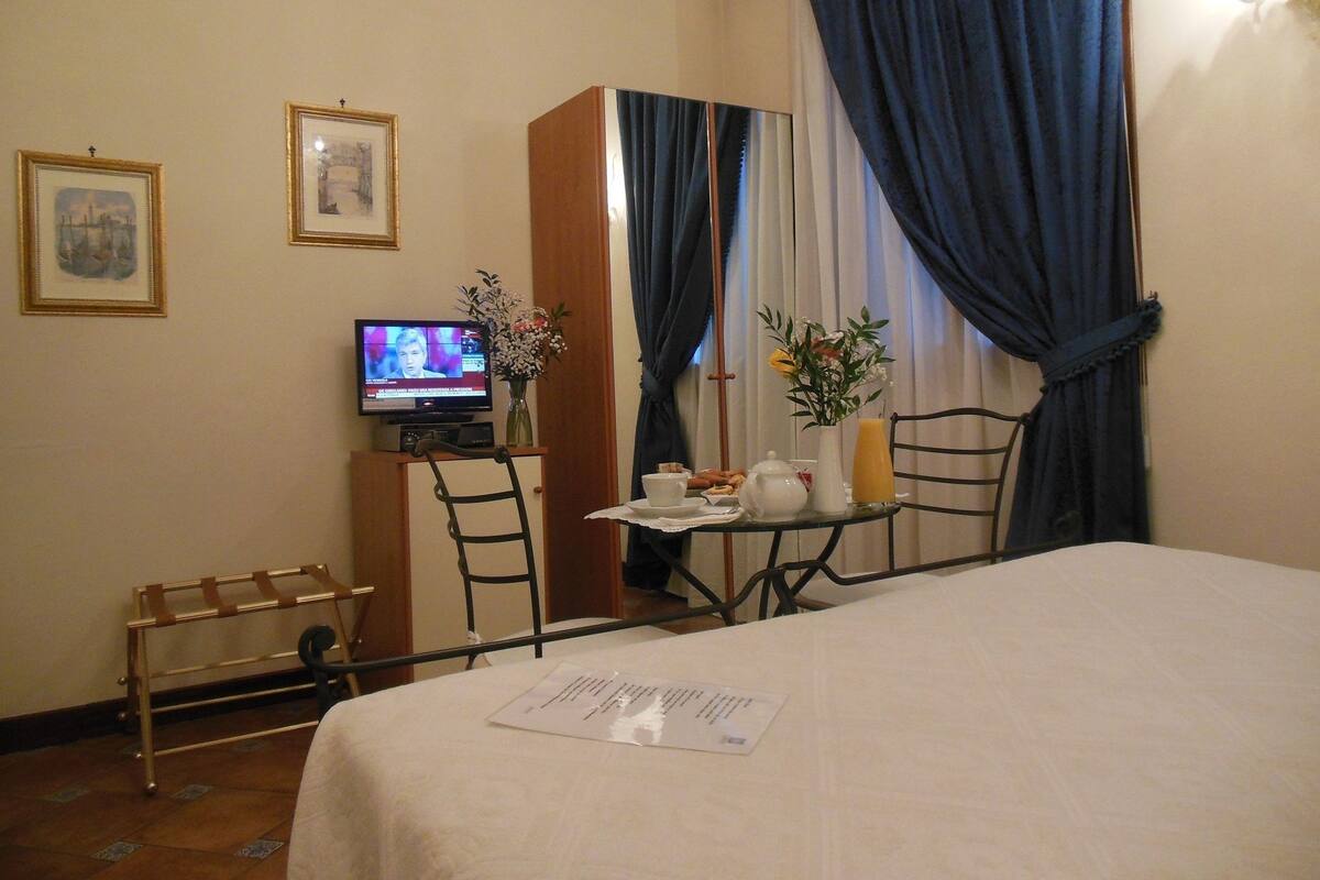 A hotel room with a bed, a small table with a vase and a teapot, a chair, a television, and a window with curtains.