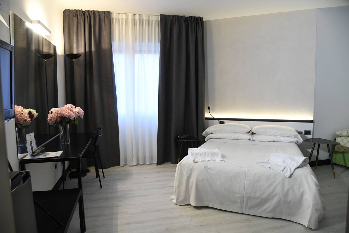 A hotel room with a bed, a desk with a chair, a mirror, and a window with curtains.