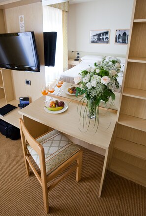 Quarto de hotel com cama, televisão, mesa de jantar com frutas e flores, e uma cadeira.