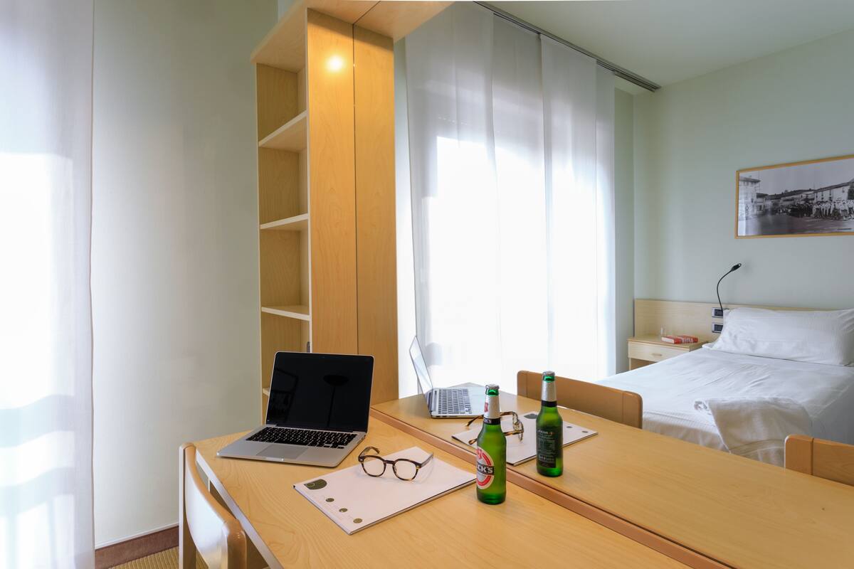 A hotel room with a wooden desk, two laptops, a pair of glasses, and a bottle of beer.