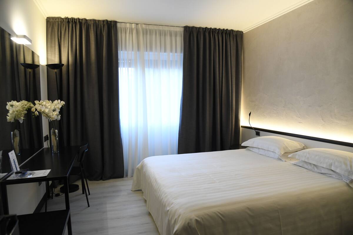A hotel room with a bed, a desk, and a chair. There is a window with curtains and a vase with flowers on the desk.