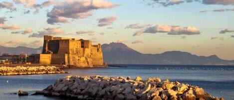 A coastal fortification with a large stone wall and a smaller building, surrounded by rocky terrain and a calm sea.
