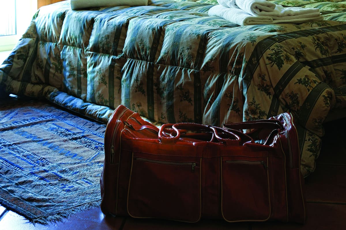 A neatly made bed with a floral comforter, a red suitcase on the floor, and a lamp on a bedside table.