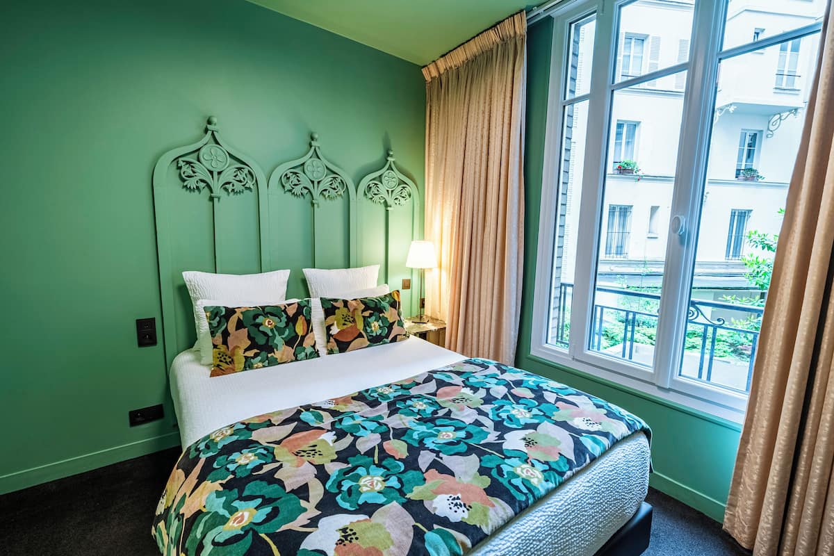 A bed with a floral bedspread, a headboard with decorative elements, a bedside lamp, and a window with a view of buildings outside.