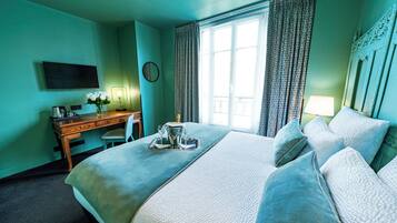 A hotel room with a large bed, a desk with a chair, a flat-screen TV, a bedside table with a lamp, and a window with curtains.