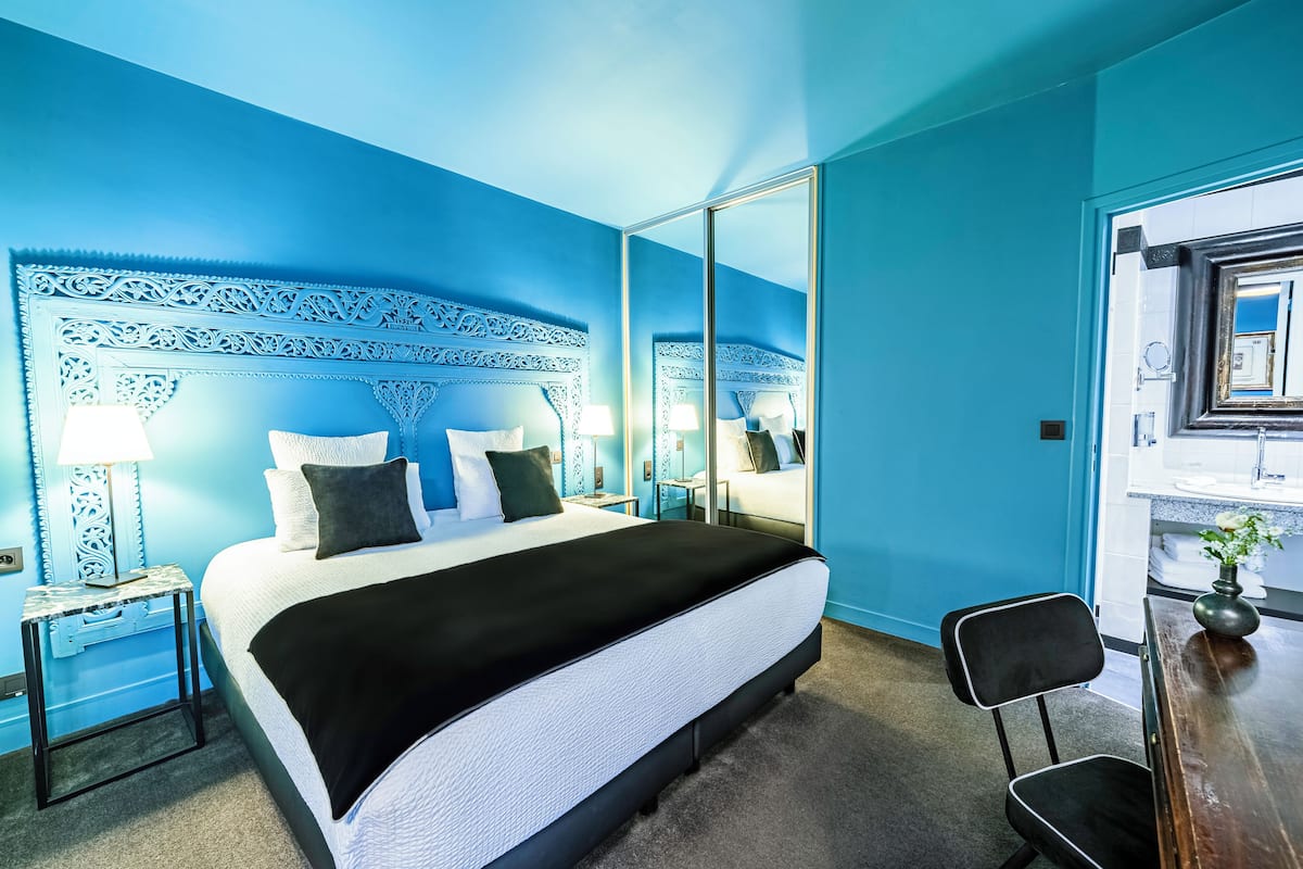 A bedroom with a large bed, a nightstand with a lamp, a chair, and a mirror.