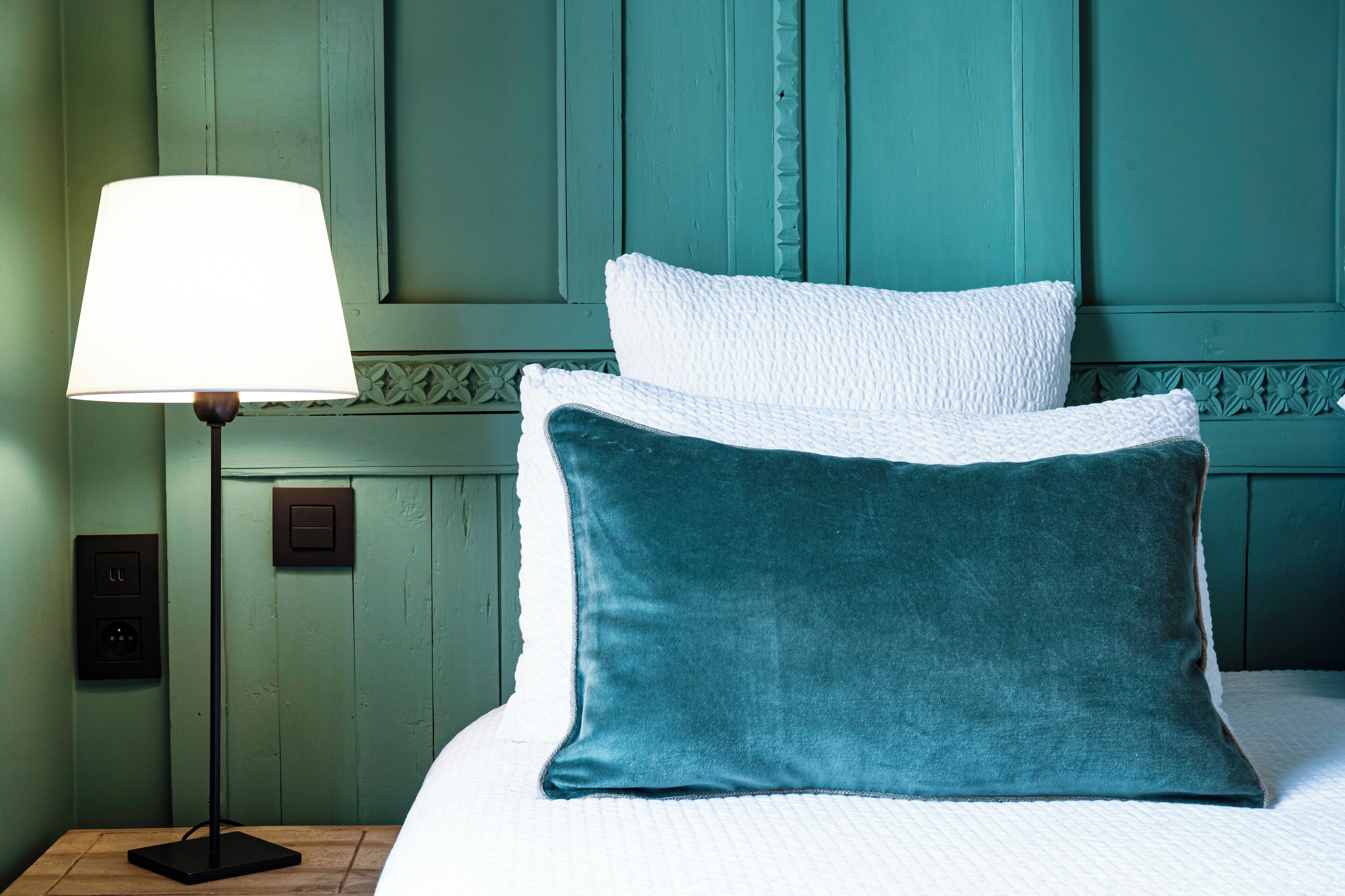 Una cama con almohadas blancas y azul turquesa, una lámpara de noche y una pared azul turquesa con un panel texturizado.