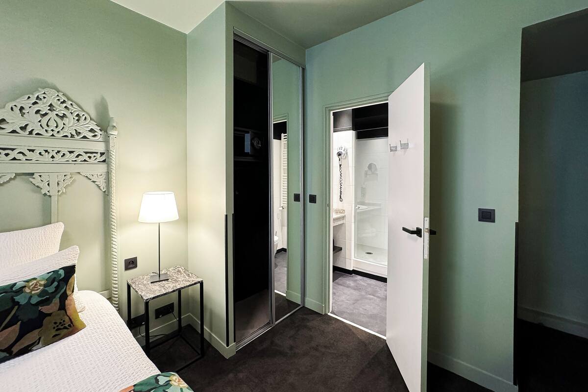 A hotel room with a bed, a bedside table with a lamp, a decorative headboard, and an open closet leading to a bathroom with a shower and toilet.