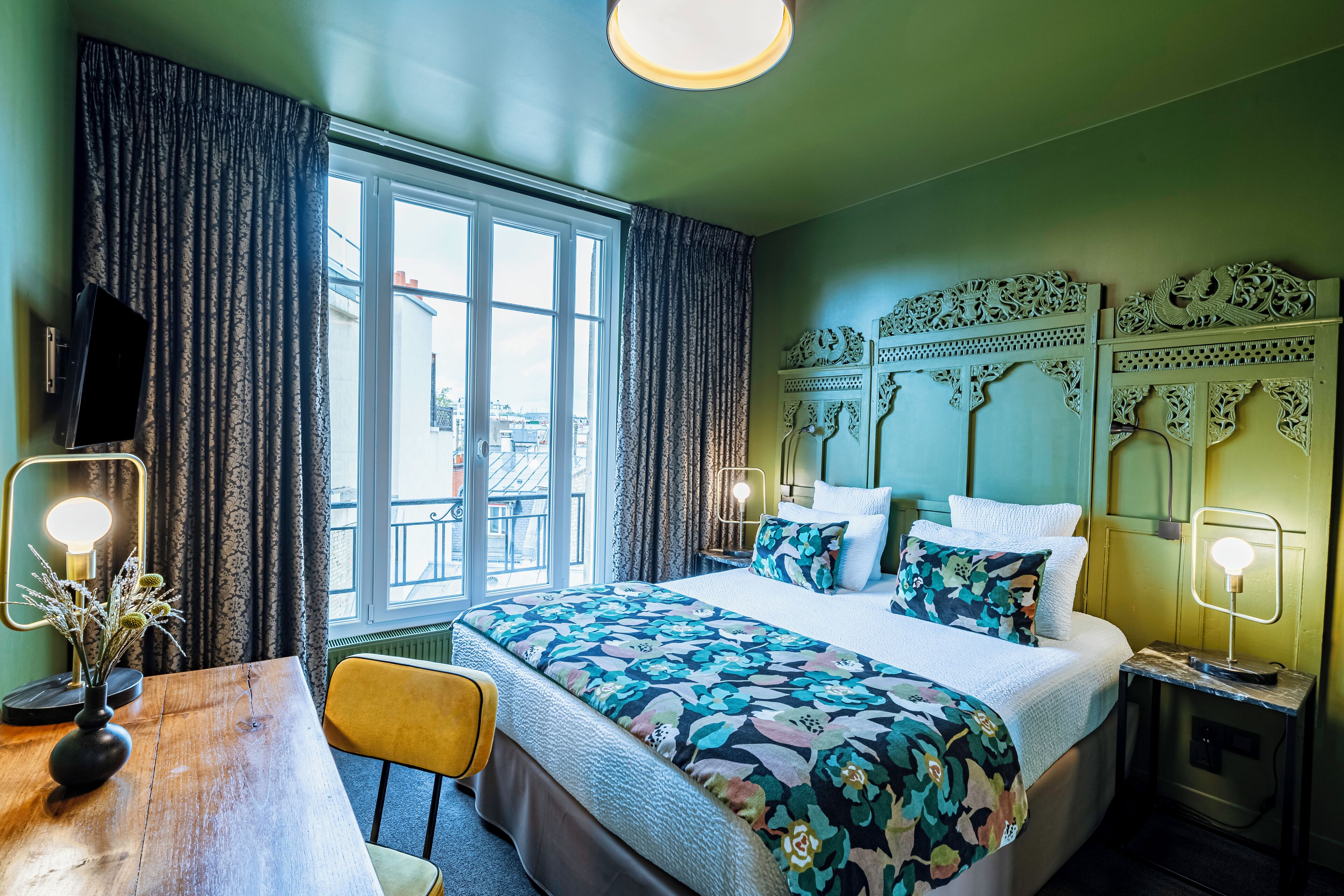 A hotel room with a large bed, a wooden desk, a chair, a TV, and a view of the city through the window.