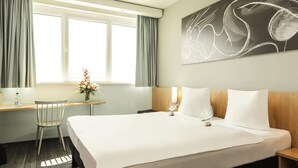 Habitación de hotel con una cama grande, un escritorio con silla, una ventana con cortinas y una obra de arte colgada en la pared.