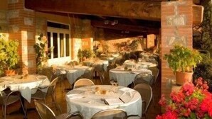Un área de comedor al aire libre con mesas redondas cubiertas con manteles blancos, rodeadas de sillas. Hay plantas y flores en macetas que aportan verdor al entorno.