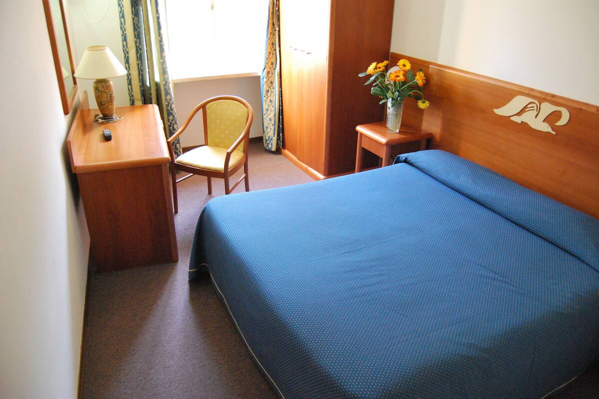 Een hotelkamer met een bed, een stoel, een bureau met een lamp en een vaas met bloemen.