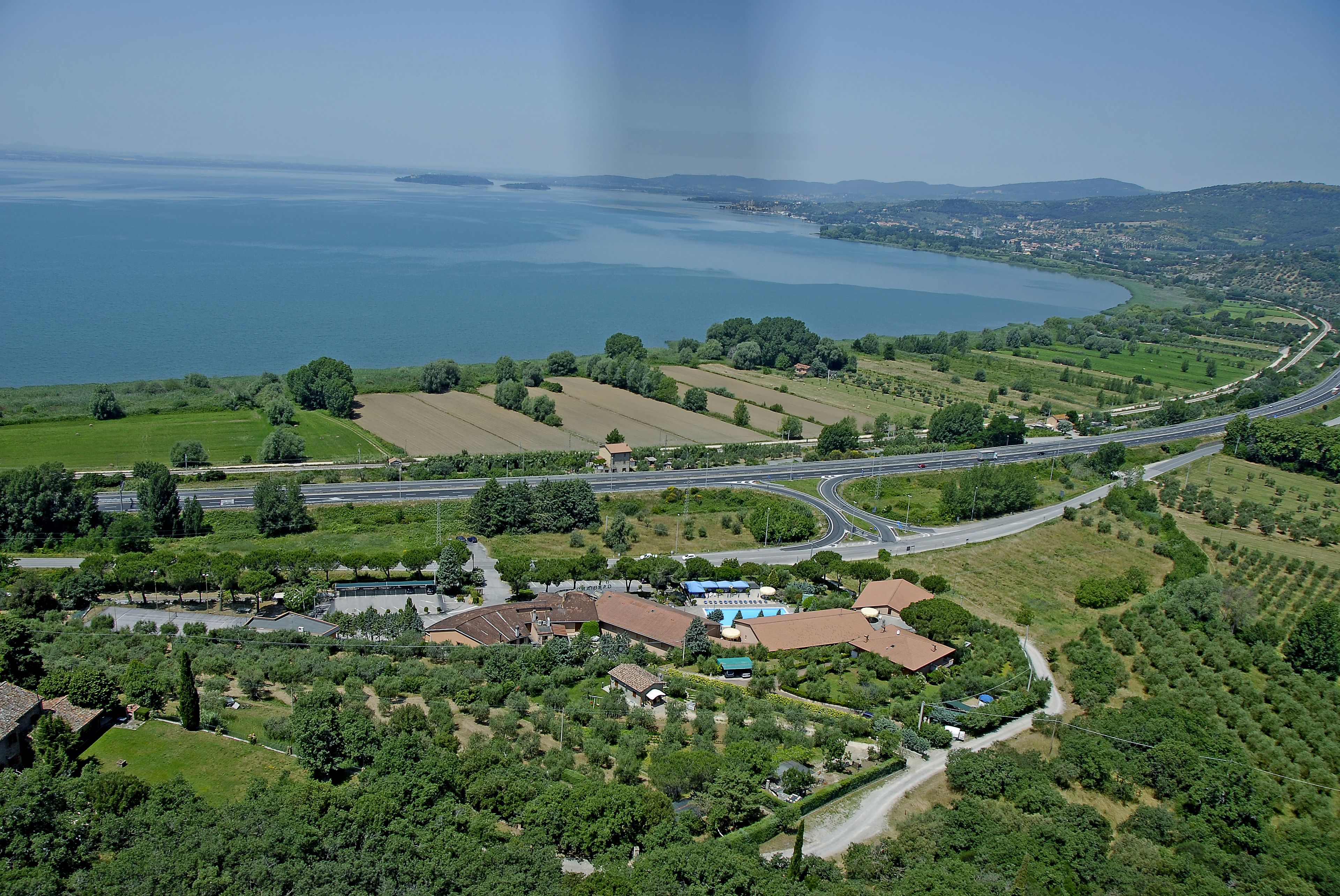 An aerial view of a coastal area with a large body of water, green fields, and a road network.