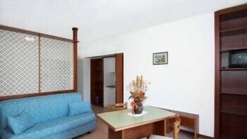 Una habitación con un sofá azul, una mesa de comedor de madera con una silla, una planta pequeña en maceta y un armario de madera.